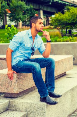 Dışarıdan Düşünen Adam. Gömlek, kot pantolon, deri ayakkabı, güneş gözlüğü, tişörte asılı, el çenesine dokunuyor, sakallı, bıyıklı genç bir adam beton bir bankta oturuyor,