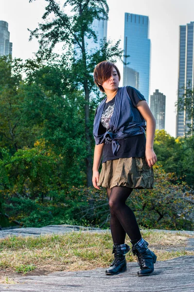 Dressing in a blue sleeveless blouse, black t shirt, green skirt,  black leggings and boots, a young Chinese girl is standing in a small words, thoughtfully looking away