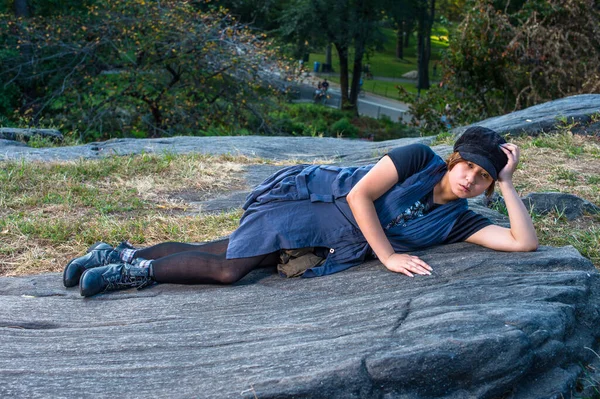 Dressing in a blue sleeveless long blouse, black leggings and boots, wearing a black corduroy cop, a young Chinese girl is lying on rocks, one hand supporting her head, into deeply thinking.