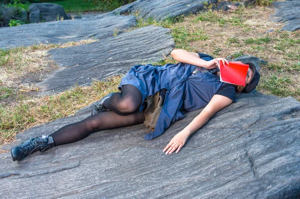 Dressing in a blue sleeveless long blouse, black leggings and boots, wearing a black corduroy cop, a young Chinese girl is lying on rocks, a red book covering her face, relaxing