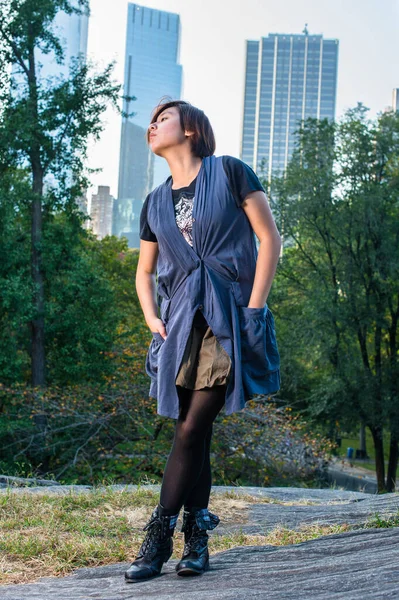 Dressing in a blue sleeveless long blouse, black leggings and boots, two hands putting in pockets, a young Chinese girl is standing in a small words.