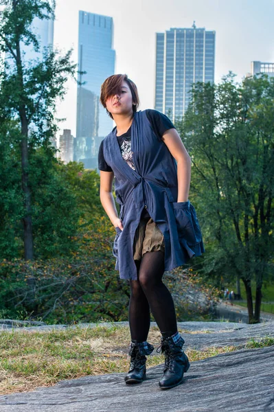 Dressing in a blue sleeveless long blouse, black leggings and boots, two hands putting in pockets, hair covering one eye, a young Chinese girl is standing in a small words. The background is high business buildings