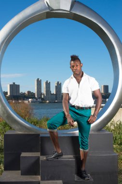 Beyaz gömlek, yeşil pantolon ve siyah deri ayakkabılar giyen, Mohawk saçlı genç siyah bir adam ringin yanında duruyor, kendine güvenen bir şekilde sana bakıyor.