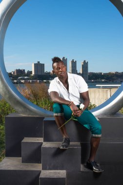 Beyaz gömlek, yeşil pantolon ve siyah deri ayakkabılar giyen, elinde beyaz bir gül tutan, Mohawk saçlı genç siyah bir adam halka yapısında seni bekliyor.