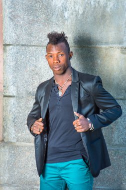 Siyah ceketli, yeşil pantolonlu, Mohawk saçlı genç siyah bir adam duvarın karşısında duruyor ve kendinden emin bir şekilde sana bakıyor.