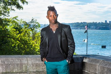 Siyah bir ceket, siyah bir atlet ve yeşil bir pantolon giymiş, iki elini cebine sokuyor, Mohawk saçlı genç bir zenci nehir kenarında duruyor, dinleniyor ve düşünüyor.