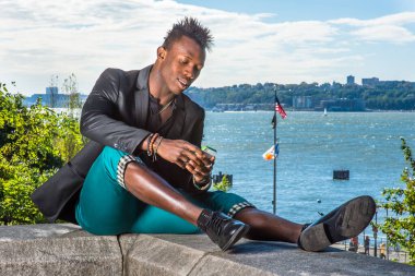 Siyah ceket, yeşil pantolon ve deri ayakkabılar giyen, Mohawk saçlı genç bir zenci nehir kenarında oturmuş, cep telefonundaki mesajları kontrol ediyor.