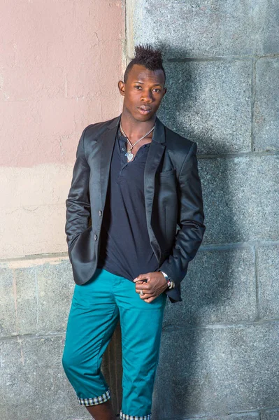 Siyah ceketli, siyah gömlekli ve yeşil pantolonlu, Mohawk saçlı genç bir adam duvarın karşısında duruyor, kendine güvenerek sana bakıyor.