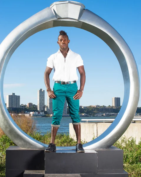 Beyaz gömlek, yeşil pantolon ve siyah deri ayakkabılar giyen, Mohawk saçlı genç siyah bir adam ringin yanında duruyor, kendine güvenen bir şekilde sana bakıyor.