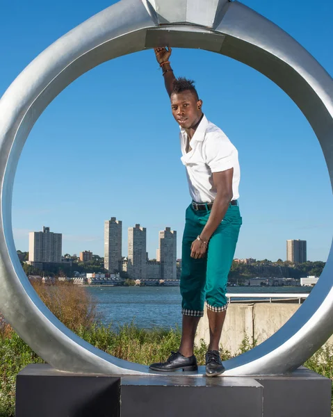 Beyaz gömlek, yeşil pantolon ve siyah deri ayakkabılar giyen, bir kolunu uzatan, Mohawk saçlı genç siyah bir adam ringin yanında duruyor, neşeyle sana bakıyor.