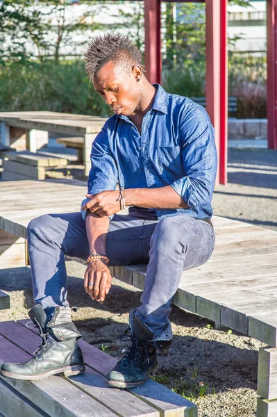 Mavi gömlekli, gri pantolonlu ve siyah deri çizmeli, Mohawk saçlı genç siyah bir adam ahşap bir yapının üzerinde oturuyor, aşağı bakıyor ve kolunun üzerinden yuvarlanıyor.