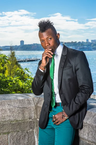 Beyaz fanilalı, siyah ceketli, yeşil pantolonlu, yeşil kravatlı, bir eli dudaklarına dokunan, Mohawk saçlı genç siyahi bir adam nehrin kenarında duruyor, dışarıda düşünüyor.