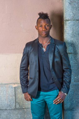 Siyah ceketli, yeşil pantolonlu, Mohawk saçlı genç siyah bir adam duvarın karşısında duruyor ve kendinden emin bir şekilde sana bakıyor.