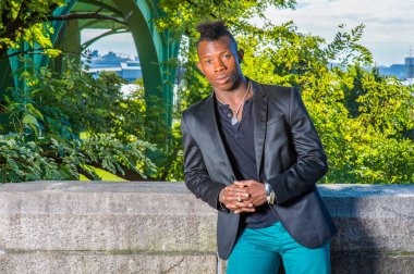 Siyah bir ceket, siyah bir atlet ve yeşil pantolon giyen, Mohawk saçlı genç bir zenci kayalık bir çitin yanında duruyor, dinleniyor.