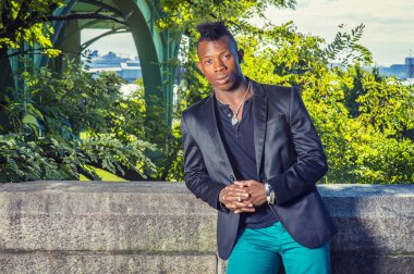 Adam dışarıda dinleniyor. Siyah bir ceket, siyah bir atlet, yeşil pantolon ve siyah saçlı genç bir adam kayalık bir çitin yanında dinleniyor. Kentsel Moda