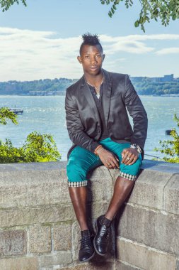 Adam dışarıda dinleniyor. Siyah bir ceket, yeşil pantolon, deri ayakkabılar giyen, Mohawk saçlı genç siyah bir adam nehir kenarındaki kayalık bir çitte oturuyor, dinleniyor. Şehir Fashio