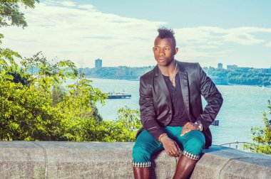 Adam dışarıda dinleniyor. Siyah ceketli, yeşil pantolonlu, Mohawk saçlı genç siyah bir adam nehir kenarındaki kayalık bir taş çitin üzerinde oturuyor, dinleniyor. Kentsel Moda