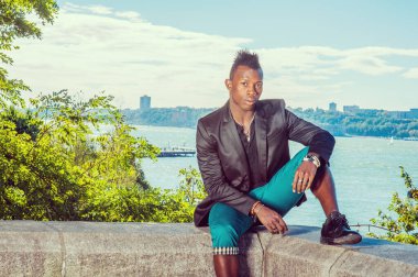 Adam dışarıda dinleniyor. Siyah ceketli, yeşil pantolonlu, Mohawk saçlı genç siyah bir adam nehir kenarındaki kayalık bir taş çitin üzerinde oturuyor, dinleniyor. Kentsel Moda