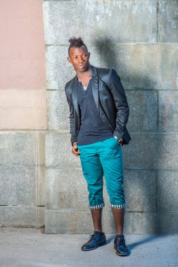 Siyah bir ceket, yeşil pantolon, deri ayakkabılar, cebinde bir el, Mohawk saçlı genç siyah bir adam duvara dayanıyor, ileriye bakıyor.