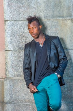 Siyah ceketli, yeşil pantolonlu, Mohawk saçlı genç siyah bir adam duvarın karşısında dikiliyor, derin derin düşünüyor.