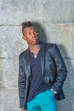 Siyah ceketli, yeşil pantolonlu, Mohawk saçlı genç siyah bir adam duvara yaslanmış, yukarı bakıyor.