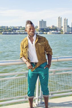 Adam dışarıda dinleniyor. Sarı yün ceket, beyaz iç çamaşırı, yeşil pantolon, cepleri dolu eller, Mohawk saçlı genç bir zenci nehir kenarında duruyor, etrafa bakıyor.