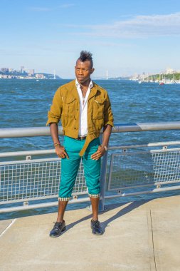 Sarı yün ceket, beyaz iç çamaşırı, yeşil pantolon, deri ayakkabılar giyen, Mohawk saçlı genç bir zenci nehir kenarında dikilip etrafa bakıyor. Arka planda bir liman ve köprü var.