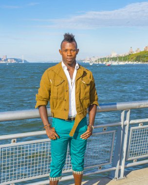 Genç adam. Sarı yünlü ceket, beyaz iç çamaşırı, yeşil pantolon giymiş mohawk saçlı genç bir zenci nehir kenarında sana bakıyor. Arka planda bir liman ve köprü var.