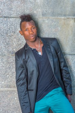 Siyah ceketli, yeşil pantolonlu, Mohawk saçlı genç siyah bir adam duvara yaslanmış, bir tarafı eğik, kendine güvenen bir şekilde sana bakıyor.