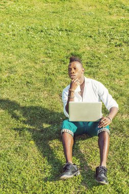 Beyaz gömlek, yeşil pantolon, deri ayakkabı, dizüstü bilgisayar ve cep telefonu tutan, çenesine dokunan, siyah saçlı genç bir adam yeşil çimlerin üzerinde oturuyor,