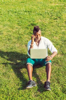 Dışarıda çalışan bir adam. Mohawk saçlı genç bir zenci yeşil çimlerin üzerinde oturuyor, dizüstü bilgisayarda çalışıyor, cep telefonuyla bir telefon görüşmesi yapıyor.