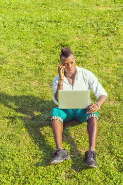 Dışarıda çalışan bir adam. Mohawk saçlı genç bir zenci yeşil çimlerin üzerinde oturuyor, dizüstü bilgisayarda çalışıyor, cep telefonuyla bir telefon görüşmesi yapıyor.