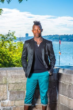 Siyah bir ceket, siyah bir atlet ve yeşil pantolon giymiş, iki elini cebine sokuyor, Mohawk saçlı genç bir zenci nehir kenarında duruyor, dinleniyor.