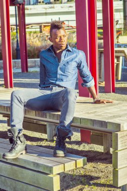 Genç işçi dışarıda dinleniyor. Mavi gömlekli, gri pantolonlu, deri çizmeli, Mohawk saçlı genç siyah bir adam ahşap bir yapının üzerinde oturuyor ve düşünüyor..