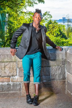 Siyah bir ceket, siyah bir atlet ve yeşil pantolon giyen, Mohawk saçlı genç bir zenci kayalık bir çitin yanında duruyor, dinleniyor.