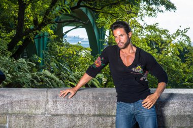 Siyah kazak ve kot pantolon giymiş, yakışıklı, seksi, orta yaşlı, bıyıklı ve sakallı bir adam yarı duvar çitinin yanında duruyor, ciddi ciddi sana bakıyor.