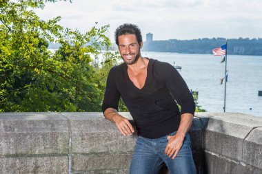 Siyah kazak ve kot pantolon giymiş, yakışıklı, seksi, bıyıklı ve sakallı orta yaşlı bir adam nehir kenarındaki yarı duvar çitlerine karşı duruyor ve sana gülümsüyor.. 