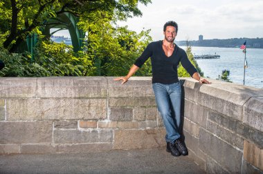 Siyah kazak, kot pantolon ve deri ayakkabılar giymiş, yakışıklı, seksi, orta yaşlı, bıyıklı ve sakallı bir adam nehir kenarındaki duvarın yarısına yaslanmış, sana gülümseyen bir şekilde bakıyor..