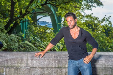 Dressing in a black sweater and blue jeans, a handsome, sexy, middle age guy with mustache and beard is standing by the half wall of fence, seriously looking at you