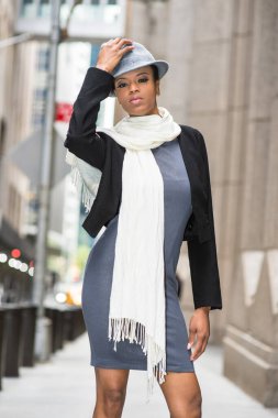 Dressing in a black woolen cropped jacket,  a gray fitted dress and a long white scarf around her neck,  a young black pretty woman is standing on the street, wearing a woolen Gangster Fedora hat