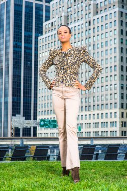 Dressing in black and yellow pattern jacket, light cream colored pants, hands on her hips, a young black businessman is standing in the front of a business district, confidently looking forward.