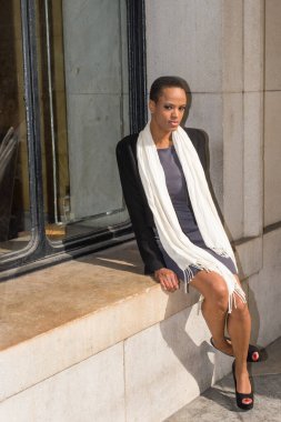 Dressing in a black jacket,  a gray fitted dress, open toe high heels and a long white scarf, a young black businesswoman is sitting on a big frame of the window outside the office to take a break.
