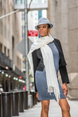 Siyah yün bir ceket, gri bir elbise, boynunda uzun beyaz bir eşarp, yün bir Gangster Fedora şapkası, genç siyah bir kadın sokakta dikiliyor.