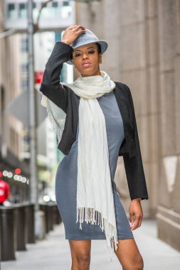 Siyah yün bir ceket, gri bir elbise, boynunda uzun beyaz bir eşarp, yün bir Gangster Fedora şapkası, genç siyah bir kadın sokakta dikiliyor..