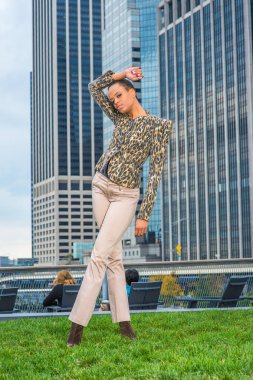 Siyah ve sarı desenli ceket, açık renkli pantolon, genç bir siyahi kadın bir iş yerinin önünde duruyor, sana bakıyor.