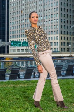 Siyah ve sarı desenli ceket, açık renkli pantolon, genç bir siyahi kadın bir iş yerinin önünde duruyor, sana bakıyor.