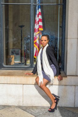 Siyah bir ceket, gri bir elbise, yüksek topuklu ayakkabılar, uzun beyaz bir eşarp, genç bir siyahi iş kadını ara vermek için ofisin dışındaki bir pencerenin çerçevesinde oturuyor.. 