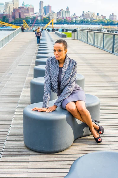 Black Woman Thinking Outside. Dressing in a gray patterned faux fur jacket, a woolen fitted dress, open toes shoes, a young professional lady is sitting on a modern style bench, relaxing, thinking, waiting for you