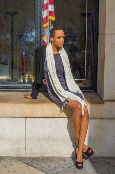 Dressing in a black jacket,  a gray fitted dress, open toe high heels, a long white scarf, a young black businesswoman is sitting on a frame of a window outside an office to take a break.