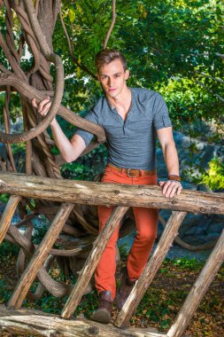 Uzun kollu yuvarlak taçlı Henley gömlek, kırmızı kot pantolon ve kahverengi çizme ayakkabı giyen genç bir adam tahta bir çitin arkasında duruyor, bir koluna dolanmış, sana bakıyor..